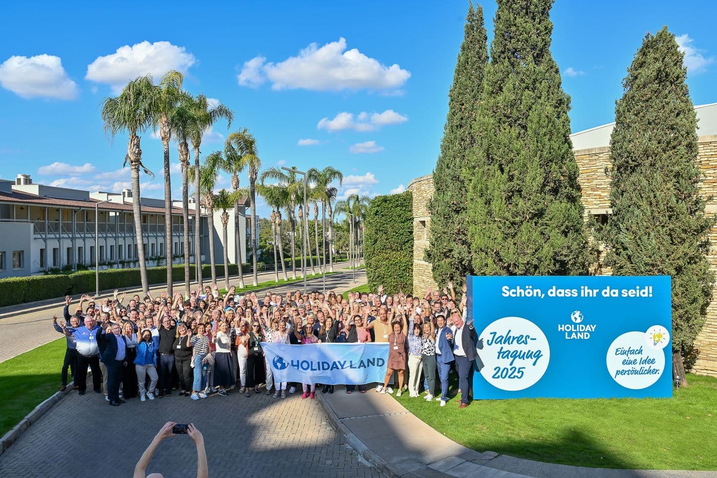 Gruppenfoto der HOLIDAY-LAND-Jahrestagung 2025 im Freien: Rund 200 Teilnehmende stehen lachend und winkend vor Palmen und Hotelgebäuden in der Lara Barut Collection in Antalya. Im Vordergrund hält eine Person ein HOLIDAY-LAND-Banner. Rechts steht ein großes blaues Willkommensschild mit den Logos von HOLIDAY LAND und dem Tagungsmotto „Einfach eine Idee persönlicher“. Der Himmel ist klar und blau.