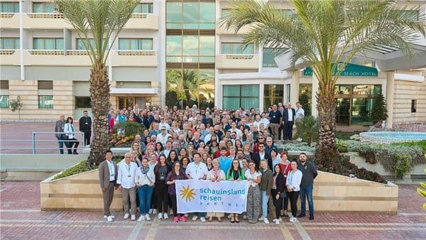Große Gruppe von Teilnehmenden der schauinsland-reisen PARTNER Jahrestagung steht vor einem Hotelgebäude mit Palmen und hält gemeinsam ein Banner mit dem Logo „schauinsland reisen PARTNER“ in die Kamera.